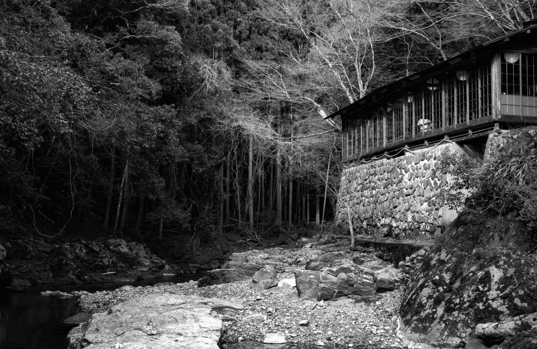 Japanese restaurant building perched over a river with a stone wall瓦そば 松右衛門, Soba Noodle Shop, Takao, Japan. 35 mm film, December 2018.